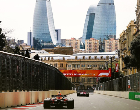 На изображении гоночные болиды движутся по городской трассе Формулы-1, окруженной защитным ограждением. На заднем плане видны современные высотные здания, сочетающиеся со старинной архитектурой