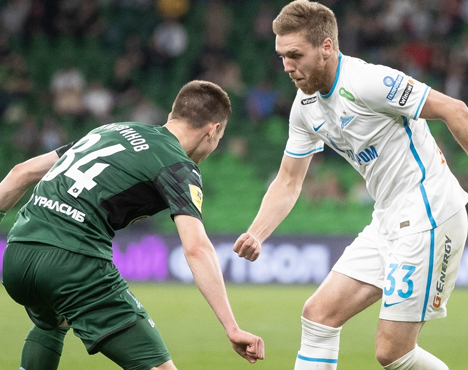 На изображении два футболиста соревнуются за мяч на поле. Игрок в тёмной форме противостоит сопернику в белой форме. Вокруг зелёная трава футбольного газона