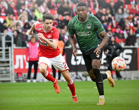 Футбольный матч: игроки в красной и зелёной форме бегут за мячом на поле. На заднем плане видно множество зрителей