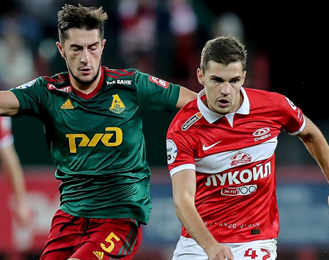 На изображении два футболиста во время матча. Один в зелёной форме с логотипом РЖД, другой в красно-белой форме с логотипом Лукойл. Они борются за мяч в динамичном эпизоде игры