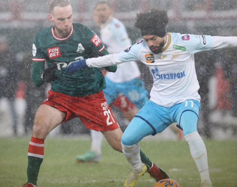 На заснеженном футбольном поле игрок в зелёной форме противостоит сопернику в бело-голубой форме. Оба стараются завладеть мячом. Видны плотная игра и зимняя погода с падающим снегом