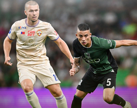 Два футболиста на поле: один в светлой форме с эмблемой и номером 11, другой в темно-зеленой форме с номером 5. Они сконцентрированно бегут, фон размыт