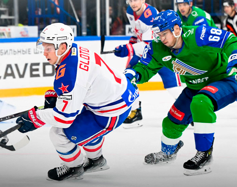 На хоккейной площадке игрок в форме с сине-красными деталями контролирует шайбу, опережая соперника в зеленой форме. Оба спортсмена сосредоточены, в окружении элементов хоккейной арены