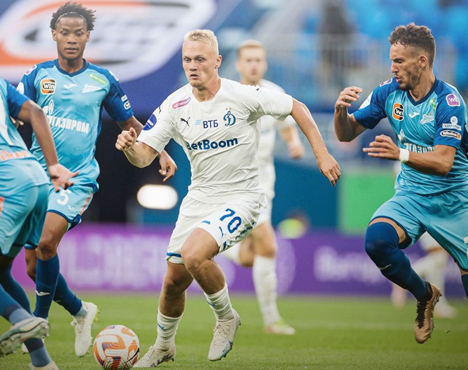 Футболисты в синих и белых формах борются за мяч на поле. Игрок в белой форме ведёт мяч, стараясь обойти соперников в голубых футболках. На заднем плане видны размытие трибун и зрители