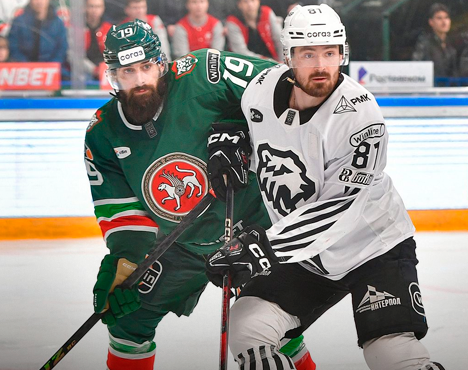 На изображении хоккеисты в момент игры. Спортсмен в зелёной форме с эмблемой включает символику с ярко-красным логотипом, а игрок в белой форме с чёрными элементами логотипа команды