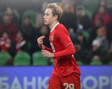 Молодой футболист в красной форме с номером 28 бежит по полю. На заднем плане видны размытые силуэты зрителей на трибунах и рекламный баннер